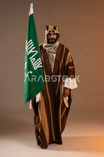 النظر إلى الكاميرا بايماءات الفخر والاعتزاز بالهوية العربية، بورتريه لرجل عربي خليجي سعودي يرتدي العقال الذهبي والدقلة والغترة، رفع علم التأسيس بتباهي، ذكرى تأسيس الدولة السعودية الأولى١٧٢٧م،يوم بدينا٢٢فبراير، صورة لكامل الجسم، خلفية بيج