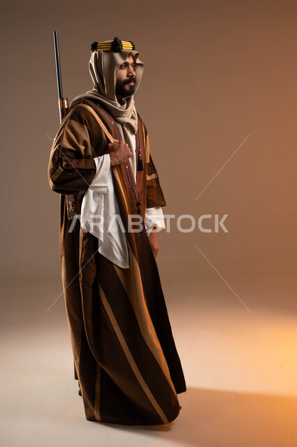 الوقوف باستقامة وشموخ، مفهوم الاصالة والعراقة عند العرب، ذكرى تأسيس الدولة الأولى 1727م، بورتريه من الجانب لشاب عربي خليجي سعودي يرتدي الدقلة والغترة والعقال الذهبي يحمل على ظهره بندقية، يوم بدينا 22 فبراير, صورة لكامل الجسم، خلفية بيج