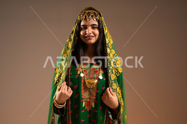 النظر إلى الكاميرا بإيماءات السعادة، العناية بالمظهر الخارجي، مفهوم التأنق والتجمل، بورتريه مقرب لإمرأة عربية خليجية سعودية ترتدي الشيلة والهامة والبشت، ارتداء أزياء المنطقة الوسطى، ذكرى تأسيس الدولة السعودية الأولى٢٢ فبراير، يوم بدينا١٧٢٧م، خلفية بيج