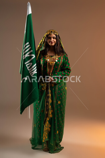 النظر الى الكاميرا بايماءات البهجة والسرور، ذكرى تأسيس الدولة السعودية الاولى ٢٢ فبراير، بورتريه لشابة عربية خليجية سعودية ترتدي الشيلة والهامة والبشت،الاحتفال بيوم بدينا ١٧٢٧م، رفع علم التأسيس بشموخ وتباهي، صورة لكامل الجسم، خلفية بيج