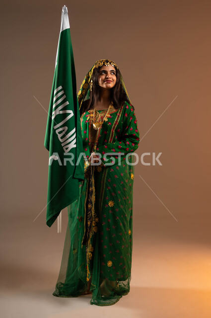 النظر إلى الأعلى بشموخ و فخر بالنتماء إلى المملكة العربية السعودية، ذكرى تأسيس الدولة السعودية الاولى ٢٢ فبراير، بورتريه لشابة عربية خليجية سعودية ترتدي الشيله والهامة والبشت تحمل بيدها علم التأسيس، الاحتفال بيوم بدينا ١٧٢٧م،صورة لكامل الجسم، خلفية بيج