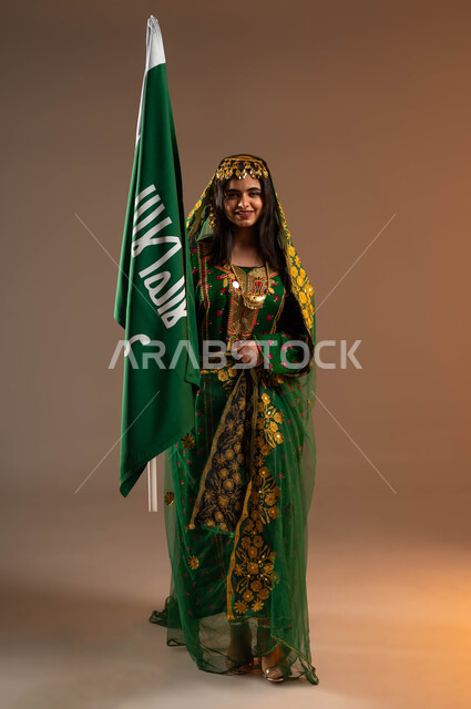 النظر الى الكاميرا بايماءات البهجة والسرور، ذكرى تأسيس الدولة السعودية الاولى ٢٢ فبراير، بورتريه لشابة عربية خليجية سعودية ترتدي الشيلة والهامة والبشت،الاحتفال بيوم بدينا ١٧٢٧م، رفع علم التأسيس بشموخ وتباهي، صورة لكامل الجسم، خلفية بيج