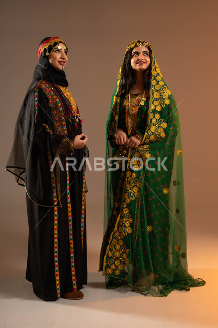 Taking care of the external appearance, the concept of femininity and elegance, a portrait of two Saudi Gulf Arab women wearing a shayla, a hama and a bisht looking somewhere, wearing the fashions of the central region, the anniversary of the founding of the first Saudi state, February 22, the day of our religion 1727 AD, a full-body portrait, beige background