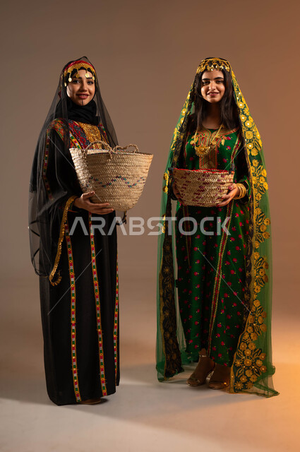مشغولات يدوية نسائية، الوقوف باستقامة والنظر الى الكاميرا بفخر، ذكرى تأسيس الدولة السعودية الاولى ٢٢ فبراير، بورتريه لشابتان عربيتان خليجيتان سعوديتان ترتديان الشيلة والهامة والبشت تحملان سلتان من القش، الاحتفال بيوم بدينا ١٧٢٧م، صورة لكامل الجسم، خلفية بيج