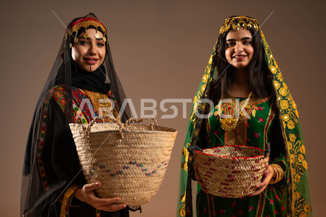 الوقوف باستقامة والنظر الى الكاميرا بفخر، ذكرى تأسيس الدولة السعودية الاولى ٢٢ فبراير، بورتريه مقرب لشابتان عربيتان خليجيتان سعوديتان ترتديان الشيلة والهامة والبشت تحملان سلتان من القش،مشغولات يدوية نسائية، الاحتفال بيوم بدينا ١٧٢٧م، خلفية بيج