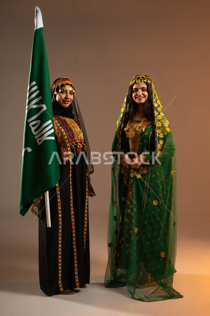 النظر الى الكاميرا بايماءات البهجة والسرور، ذكرى تأسيس الدولة السعودية الاولى ٢٢ فبراير، بورتريه لشابتان عربيتان خليجيتان سعوديتان ترتديان ازياء المنطقة الوسطى ،الاحتفال بيوم بدينا ١٧٢٧م، رفع علم التأسيس بشموخ وتباهي، صورة لكامل الجسم، خلفية بيج