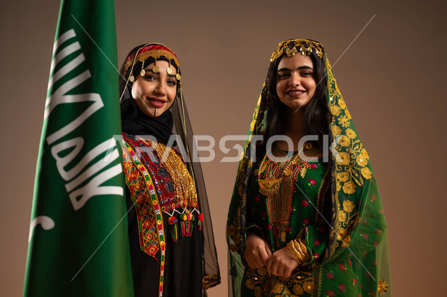 ذكرى تأسيس الدولة السعودية الاولى ٢٢ فبراير، بورتريه مقرب لشابتان عربيتان خليجيتان سعوديتان ترتديان أزياء المنطقة الوسطى،الاحتفال بيوم بدينا ١٧٢٧م، اناقة الازياء الشعبية في المناسبات الوطنية، رفع علم التأسيس بشموخ وتباهي، خلفية بيج