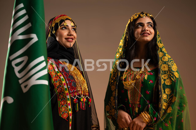 ارتداء الازياء الشعبية في المناسبات الوطنية، رفع علم التأسيس بشموخ وتباهي، الاحتفال بذكرى تأسيس الدولة السعودية الاولى ٢٢ فبراير، بورتريه مقرب لشابتان عربيتان خليجيتان سعوديتان ترتديان أزياء المنطقة الوسطى، يوم بدينا ١٧٢٧م، خلفية بيج