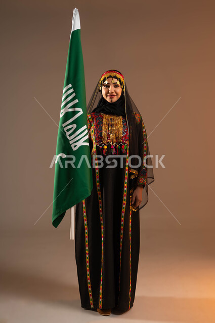 الوقوف بفخر واعتزاز، إيماءات الفرح والسرور احتفالا بذكرى تأسيس الدولة السعودية الاولى ٢٢ فبراير، بورتريه لامرأة عربية خليجية سعودية ترتدي زي المنطقة الوسطى تمسك بيدها علم التأسيس، يوم بدينا ١٧٢٧م، أزياء شعبية تقليدية، صورة لكامل الجسم