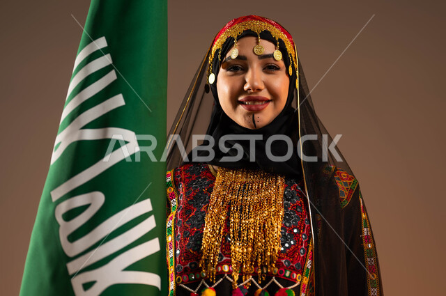 Looking at the camera with gestures of joy and pleasure, wearing popular costumes on national occasions, raising the founding flag proudly and proudly, celebrating the anniversary of the founding of the first Saudi state on February 22, a close-up portrait of a young Saudi Gulf Arab woman wearing the uniform of the central region, Day of Dina 1727 AD, beige background.