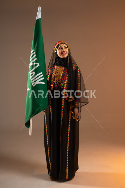 النظر الى السماء بنظرات النصر والبهجة، إيماءات الفرح والسرور احتفالا بذكرى تأسيس الدولة السعودية الاولى ٢٢ فبراير، بورتريه لشابة عربية خليجية سعودية ترتدي زي المنطقة الوسطى تمسك بيدها علم التأسيس، يوم بدينا ١٧٢٧م، أزياء شعبية تقليدية، صورة لكامل الجسم