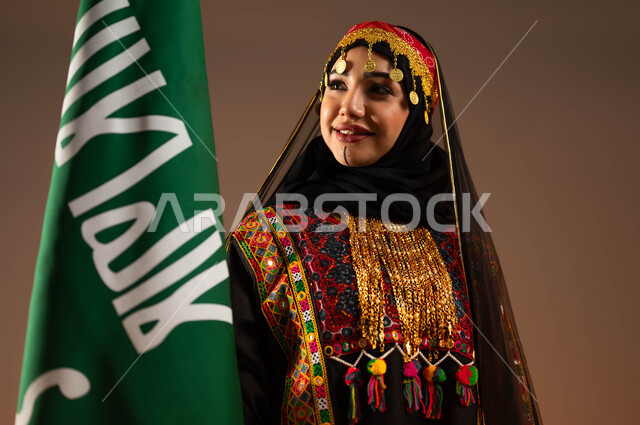 رفع علم التأسيس بشموخ وتباهي، الاحتفال بذكرى تأسيس الدولة السعودية الاولى ٢٢ فبراير، بورتريه مقرب لشابة عربية خليجية سعودية ترتدي الشيلة والبشت والهامة، ارتداء الازياء الشعبية في المناسبات الوطنية، يوم بدينا ١٧٢٧م، ايماءات البهجة والسعادة، خلفية بيج
