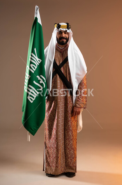 إحياء التراث الشعبي في المناسبات الوطنية, بورتريه لشاب عربي خليجي سعودي مبتسم يرتدي أزياء المنطقة الوسطى يمسك بيده علم التأسيس باعتزاز وينظر إلى الكاميرا, يوم بدينا 1727م, ذكرى تأسيس الدولة السعودية الأولى في 22 فبراير, صورة الجسم بالطول الكامل, خلفية بيج
