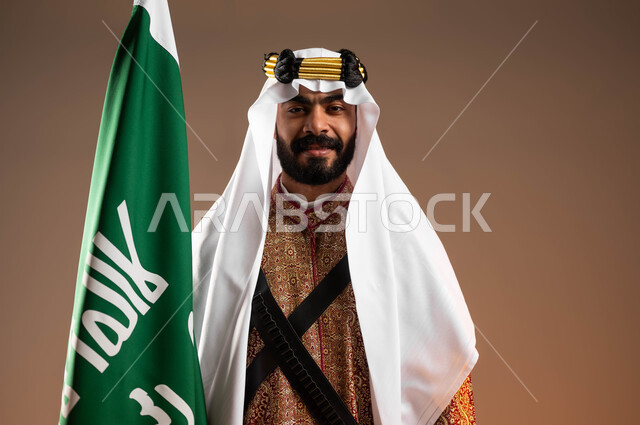 النظر إلى الكاميرا بثقة وفخر, بورتريه مقرب لشاب عربي خليجي سعودي مبتسم  يرتدي أزياء المنطقة الوسطى يمسك بيده علم التأسيس باعتزاز, يوم بدينا 1727م, ذكرى تأسيس الدولة السعودية الأولى في 22 فبراير, إحياء التراث الشعبي في المناسبات الوطنية, خلفية بيج