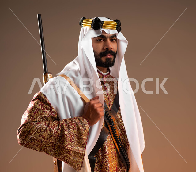 ذكرى تأسيس الدولة السعودية الأولى في 22 فبراير, ارتداء أزياء المنطقة الوسطى في المناسبات الوطنية، إحياء التراث الخليجي, بورتريه مقرب لشاب عربي خليجي سعودي يرتدي الدقلة والغترة والعقال الذهبي يمسك بيده البندقية وينظر إلى الكاميرا, يوم بدينا 1727م, خلفية بيج