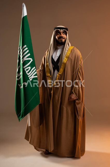 ذكرى تأسيس الدولة السعودية الأولى في 22 فبراير, رفع علم يوم التأسيس باعتزاز, بورتريه لشاب عربي خليجي سعودي يرتدي المحزم والجبة والعقال ينظر إلى شيء ما, يوم بدينا 1727م, ارتداء أزياء المنطقة الجنوبية في المناسبات الوطنية, صورة الجسم بالطول الكامل. خلفية بيج