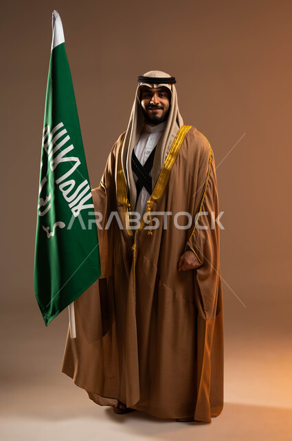 رفع علم يوم التأسيس باعتزاز, بورتريه لشاب عربي خليجي سعودي يرتدي المحزم والجبة والعقال ينظر إلى الكاميرا, يوم بدينا 1727م, ذكرى تأسيس الدولة السعودية الأولى في 22 فبراير, ارتداء أزياء المنطقة الجنوبية في المناسبات الوطنية, صورة الجسم بالطول الكامل. خلفية بيج