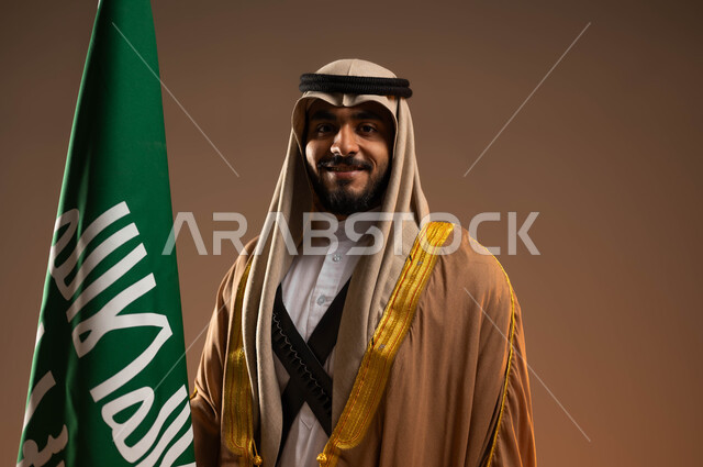 النظر إلى الكاميرا بإيماءات الثقة والفخر, بورتريه مقرب لشاب عربي خليجي سعودي يرتدي المحزم والجبة والعقال يمسك علم التأسيس باحترام, ذكرى تأسيس الدولة السعودية الأولى في 22 فبراير, يوم بدينا 1727م, ارتداء أزياء المنطقة الجنوبية في المناسبات الوطنية, خلفية بيج