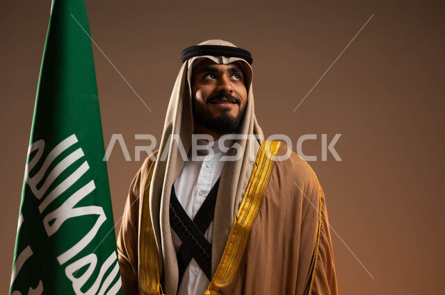 التأمل والنظر إلى شيء ما, بورتريه مقرب لشاب عربي خليجي سعودي يرتدي المحزم والجبة والعقال يمسك علم التأسيس بفخر, ذكرى تأسيس الدولة السعودية الأولى في 22 فبراير, يوم بدينا 1727م, ارتداء أزياء المنطقة الجنوبية في المناسبات الوطنية, خلفية بيج