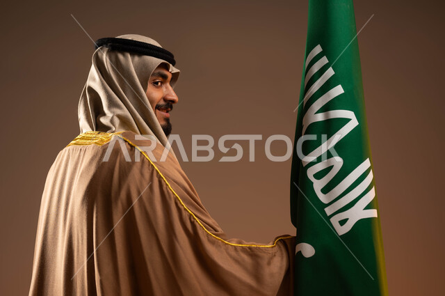 ارتداء الأزياء الشعبية التقليدية في المناسبات الوطنية, التباهي بإنجازات البلاد, بورتريه مقرب من الخلف لرجل عربي خليجي سعودي مبتسم يرتدي البشت والعقال يمسك علم التأسيس بفخر, يوم بدينا 1727م, ذكرى تأسيس الدولة السعودية الأولى في 22 فبراير, خلفية بيج