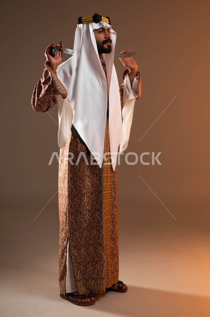 النظر إلى شيء ما بفخر واعتزاز, بورتريه لرجل عربي خليجي سعودي يرتدي الغترة والعقال الذهبي والدقلة يمسك بيده السيف, ارتداء أزياء المنطقة الوسطى في المناسبات الوطنية, يوم بدينا 1727م, ذكرى تأسيس الدولة السعودية الأولى في 22 فبراير, صورة الجسم بالكامل, خلفية بيج