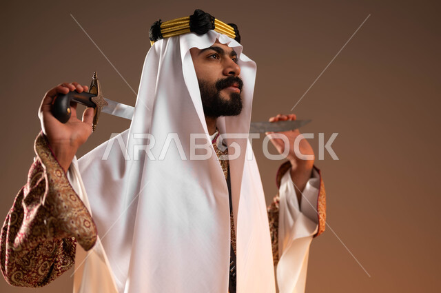 Contemplating, thinking and looking at something, a close-up portrait of a Saudi Gulf Arab man wearing a ghutra, a golden aqal and a dakhla holding a sword in his hand, wearing the costumes of the central region on national occasions, Day of Our Day 1727 AD, the anniversary of the founding of the first Saudi state on February 22, beige background