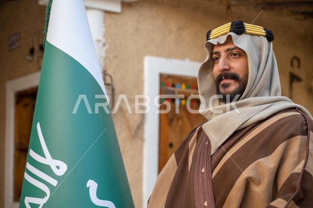 Anniversary of the founding of the first Saudi state on February 22, standing proudly in national identity, close-up photo of a Saudi Gulf Arab man wearing a bisht and aqal holding the founding flag proudly and looking at something, Day of Dina 1727 AD, old mud heritage houses