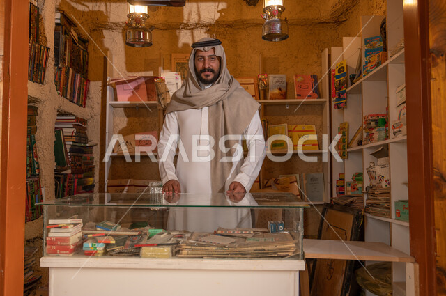 ذكرى تأسيس الدولة السعودية الأولى 22 فبراير، يوم بدينا 1727 م، المكتبات القديمة في الأحياء التراثية الطينية، النظر إلى الكاميرا بإيماءات السعادة, أماكن بيع الكتب التاريخية والمستلزمات المكتبية, شاب عربي خليجي سعودي مبتسم يرتدي الزي التقليدي يقف داخل مكتبته