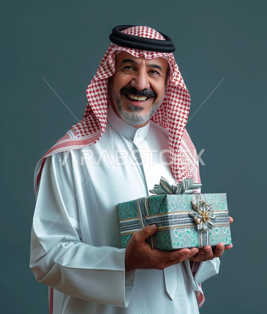 إهداء وتقديم الهدايا الفاخرة، أعياد ومناسبات ومفاجآت سعيدة, بورتريه لرجل عربي خليجي سعودي مبتسم يرتدي الثوب التقليدي يحمل بيديه علبة هدايا مغلفة ينظر الى الكاميرا بإيماءات السرور، خلفية رمادية