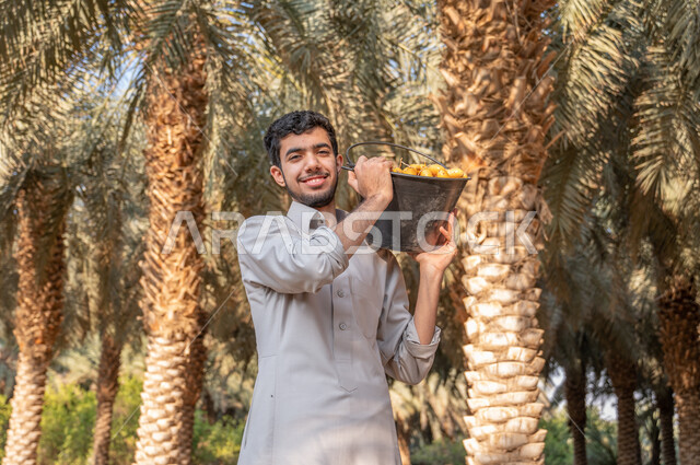 النظر الى الكاميرا بايماءات السرور، شاب عربي خليجي سعودي يرتدي الزي الشعبي يحمل على كتفه دلو مليء بالرطب، موسم الحصاد وجني الثمار في مزارع النخيل، اتقان مهنة الزراعه والفلاحة، دور الفلاحين السعوديين في رعاية الاراضي الزراعية، منتجات ومحاصيل وطنية محلية
