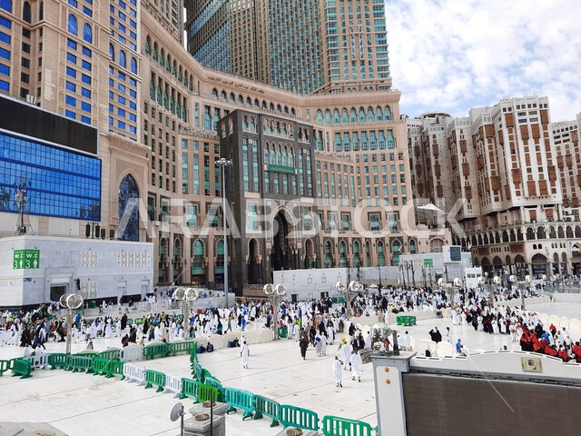 وقوف الحجاج والمعتمرين في الساحة الخارجية للحرم المكي الشريف في مكة المكرمه، أماكن ومعالم دينية اسلامية مقدسة, التجمع الاسلامي للعبادة والتقرب إلى الله, الفنادق والأبراج الفاخرة المحيطة بالمسجد الحرام, منظر السماء الزرقاء المليئة بالغيوم، أداء مناسك الحج والعمرة