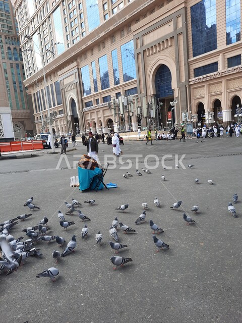 طيور الحمام تأكل الحبوب في الفناء الخارجي للحرم المكي في مكة المكرمه، الفنادق والأبراج الفاخرة المحيطة بالمسجد,  قدوم الحجاج والمعتمرين من كل انحاء العالم لأداء العبادات والتقرب الى الله, أماكن ومعالم دينية اسلامية مقدسة, السماء الزرقاء المليئة بالغيوم