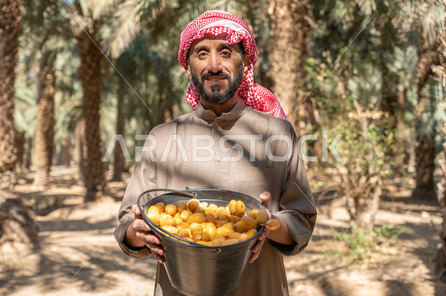 اتقان مهنة الزراعه، النظر الى الكاميرا بايماءات السرور، صورة مقربة لفلاح عربي خليجي سعودي يرتدي الزي الشعبي يحمل بيده دلو مليء بالرطب، موسم الحصاد وجني الثمار في مزارع النخيل، دور الفلاحين السعوديين في رعاية الاراضي الزراعية، منتجات ومحاصيل وطنية محلية