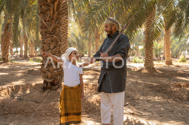 محاصيل زراعيه محليه، الاستعداد لحراثة وحفر الارض الزراعيه، ولد عربي خليجي سعودي يرتدي الزي الشعبي التقليدي يشير بيده الى شيء ما ويقف مع والده داخل مزارع النخيل المثمرة في المملكة، مفهوم مهنة الزراعة والفلاحة، الحفاظ على تراث زراعي غني
