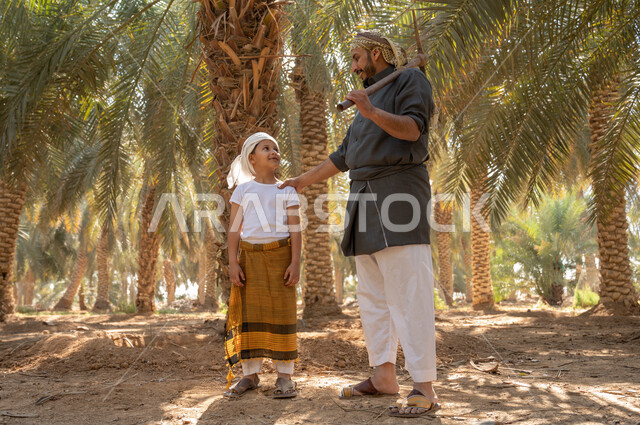 Preparing to plow and dig agricultural land, a Saudi Gulf Arab man wearing a traditional folk costume holding a digging tool in his hand, standing with his son inside the fruitful palm farms in the Kingdom, the concept of the profession of agriculture and agriculture, local agricultural crops, preserving a rich agricultural heritage