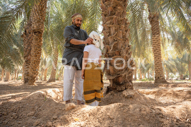 اتقان مهنة الفلاحة والزراعه ،تربية الأبناء على حب المهن الشعبيه ،فلاح عربي خليجي سعودي يرتدي الزي الشعبي يقوم بتعديل اللباس لابنه و ينظر اليه بإيماءات السرور في أحد مزارع النخيل، مفهوم العائلة السعيدة ،المحافظة على التراث الزراعي الغني، البيئة والانتاج المحلي