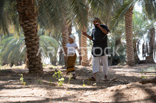 Pointing to something, local agricultural crops, the concept of the profession of agriculture and cultivation, preserving a rich agricultural heritage, preparing to plow the agricultural land, a Saudi Arabian Gulf farmer wearing a traditional folk costume, holding a digging tool in his hand, walking around with his son inside the fruitful palm farms with gestures of pleasure.