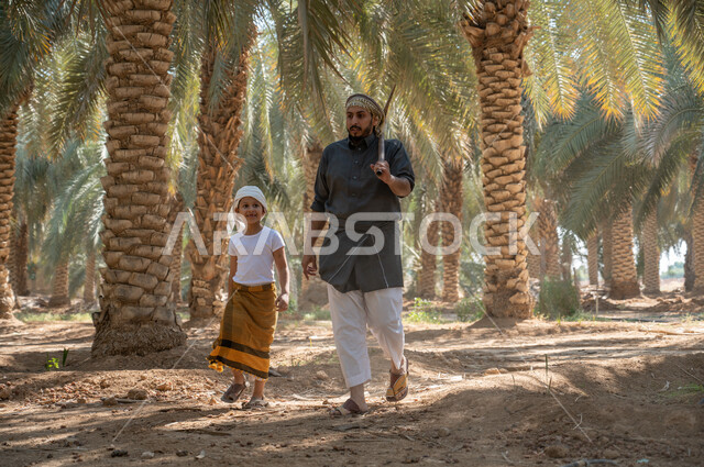 Interest in planting palm trees, preserving a rich agricultural heritage, preparing to plow the agricultural land, a Saudi Arabian Gulf farmer wearing a traditional folk costume, holding a drilling tool in his hand, walking around with his son inside the fruitful palm farms with gestures of pleasure, local agricultural crops.
