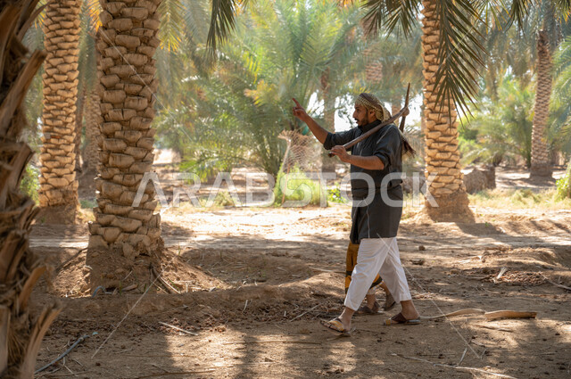 الاشارة الى شيء ما، محاصيل محليه زراعية ،مفهوم مهنة الزراعة والفلاحة، الحفاظ على تراث زراعي غني ،الاستعداد لحراثة الارض الزراعيه، فلاح عربي خليجي سعودي يرتدي الزي الشعبي التقليدي يحمل بيده أداة الحفر يتجول مع ابنه داخل مزارع النخيل المثمرة بإيماءات السرور