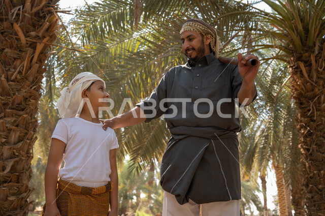 The concept of the profession of agriculture and farming, preserving a rich agricultural heritage, preparing to plow the agricultural land, a close-up picture of a Saudi Arabian Gulf farmer wearing a traditional folk costume, holding a drilling tool in his hand, standing with his son inside the fruitful palm farms with gestures of pleasure, local agricultural crops.