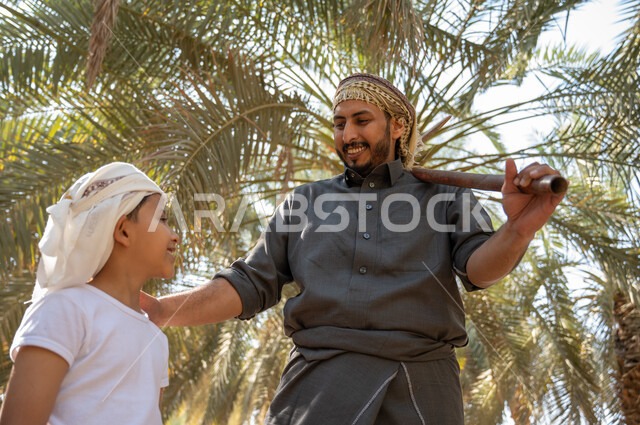 الحفاظ على تراث زراعي غني ،الاستعداد لحراثة الارض الزراعيه، صورة مقربه لفلاح عربي خليجي سعودي يرتدي الزي الشعبي التقليدي يحمل بيده أداة الحفر يقف مع ابنه داخل مزارع النخيل المثمرة بإيماءات السرور، مفهوم مهنة الزراعة والفلاحة، محاصيل زراعيه محليه