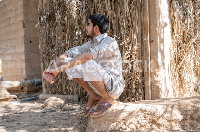 النظر الى شيء ما بإيماءات شرود الذهن، شاب عربي خليجي سعودي يرتدي الزي الشعبي ويجلس في أحد مزارع النخيل في المملكة العربيه السعودية، التنزه في أوقات الفراغ، مفهوم حياة البدو الشعبيه ،الاهتمام في زراعه اشجار النخيل الخضراء