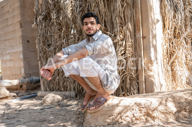 التنزه في أوقات الفراغ، النظر الى الكاميرا بإيماءات السرور، شاب عربي خليجي سعودي يرتدي الزي الشعبي ويجلس في أحد مزارع النخيل في المملكة العربيه السعودية، مفهوم حياة البدو الشعبيه ،الاهتمام في زراعه اشجار النخيل الخضراء