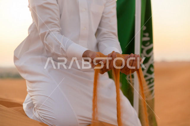 رجل سعودي بجانبه علم السعودية ، يمسك الرمال الذهبية بيديه فوق تلال احدى المناطق الصحراوية بالمملكة العربية السعودية ، اليوم الوطني السعودي ، اليوم الوطني ، مناطق صحراوية ، جمال الطبيعة السعودية