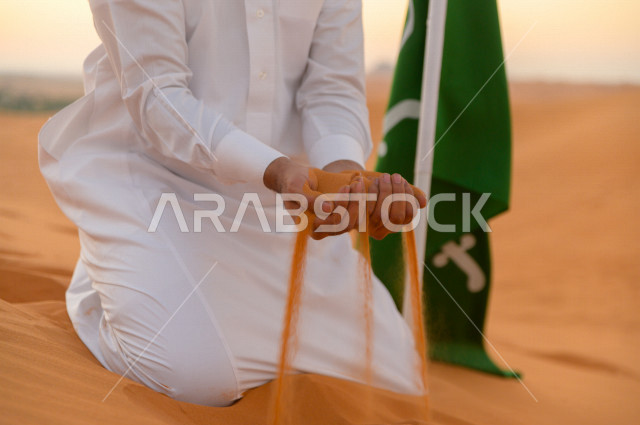 A Saudi man next to the Saudi flag, holding golden sand in his hands over the hills of one of the desert regions in the Kingdom of Saudi Arabia, Saudi National Day, National Day, desert areas, the beauty of Saudi nature