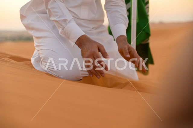 رجل سعودي بجانبه علم السعودية ، يمسك الرمال الذهبية بيديه فوق تلال احدى المناطق الصحراوية بالمملكة العربية السعودية ، اليوم الوطني السعودي ، اليوم الوطني ، مناطق صحراوية ، جمال الطبيعة السعودية