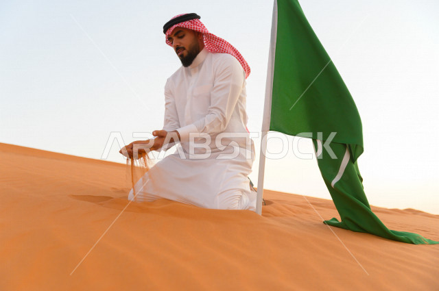 رجل سعودي بجانبه علم السعودية ، يمسك الرمال الذهبية بيديه فوق تلال احدى المناطق الصحراوية بالمملكة العربية السعودية ، اليوم الوطني السعودي ، اليوم الوطني ، مناطق صحراوية ، جمال الطبيعة السعودية