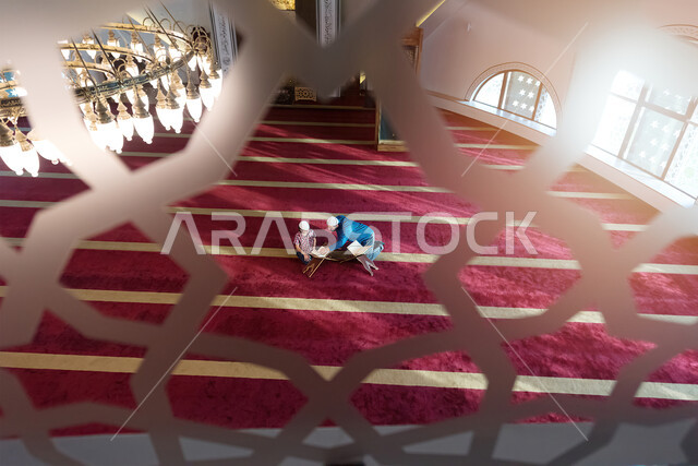 A Saudi Muslim man sits inside the mosque with his little child next to him, reading the Holy Qur’an, memorizing the Holy Qur’an for children, worshiping and getting closer to God.