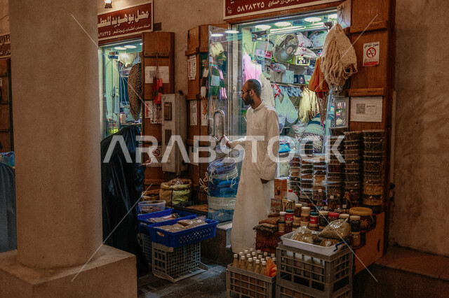 رجل عربي خليجي سعودي يرتدي الثوب التقليدي الأبيض يقف أمام متجره يقوم باداء عمله، سوق القيصرية في مدينة الاحساء، أماكن وأسواق شعبية تراثية مشهورة