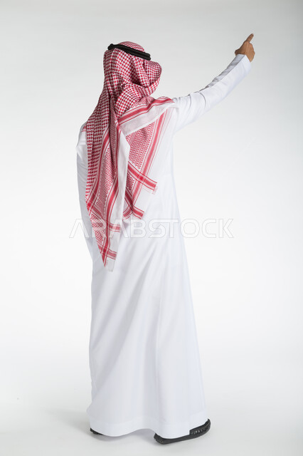 The concept of masculinity and self-confidence, elegance and attention to external appearance, a portrait from the back of a Saudi Gulf Arab man wearing a shemagh and traditional dress, standing straight and pointing with his fingers at something, full-length body portrait, white backgroun